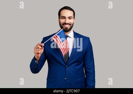 Porträt eines positiv optimistischen bärtigen Mannes mit Flagge der vereinigten Staaten von amerika, Patriotismus, Unabhängigkeit und offiziellem Anzug. Innenaufnahme des Studios isoliert auf grauem Hintergrund. Stockfoto