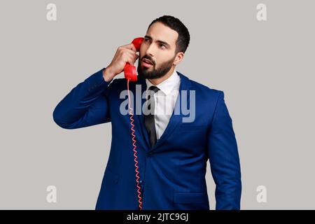 Nachdenklicher bärtiger Mann, der Telefonhörer hält, Gespräche führt, mit nachdenklicher Ausdrucksweise wegschaut und einen offiziellen Style-Anzug trägt. Innenaufnahme des Studios isoliert auf grauem Hintergrund. Stockfoto