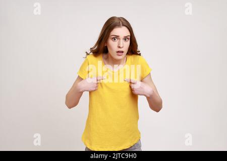 Das bin ich. Staunend attraktives Teenager-Mädchen mit braunen Haaren zeigt sich mit wundertem Ausdruck, überrascht von eigenem Erfolg, Unglaube Reaktion. Innenaufnahme des Studios isoliert auf grauem Hintergrund. Stockfoto