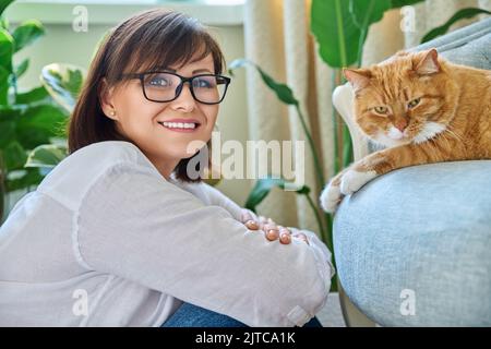 Porträt einer lächelnden Frau mit einer Katze, die auf einem Sessel zu Hause liegt Stockfoto