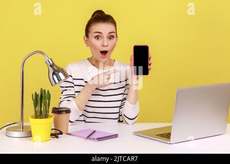 Schockiert erstaunt Frau zeigt auf Smartphone mit leerem weißen Display, überrascht von neuen Modell Blick auf die Kamera mit offenem Mund. Studio-Innenaufnahme isoliert auf gelbem Hintergrund. Stockfoto