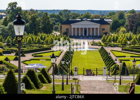 Vom Schlossberg aus bietet diese Aussicht über den Botanischen Garten der Universität Uppsala (Botaniska trädgården, auch bekannt als der Schlosspark) mit seinem Brunnen und dem Orangerie dahinter, Schweden. Weiße Zelte und Tische im Garten deuten darauf hin, dass eine Veranstaltung stattfindet. Der Architekt Carl Hårleman entwarf den barocken Garten, der 1655 gegründet und 1787 hier an den Fuß des Uppsala-Schlosses verlegt wurde. Eine Reihe der pyramidalen Hecken zeigt einen Makel. Die Orangerie, „Linneanum“, wurde nach 1787 als Geschenk von König Gustav III. erbaut. Der Garten dient noch heute der Forschung und Lehre der Universität und zieht mehr Stockfoto