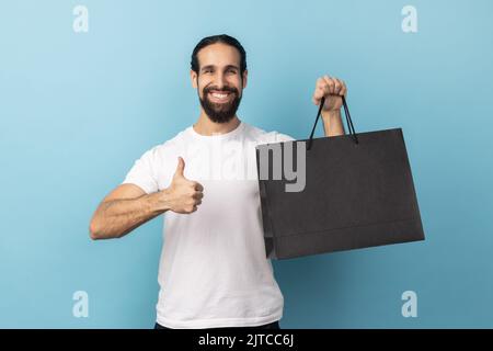 Porträt eines Mannes mit Bart, der weißes T-Shirt trägt und Daumen nach oben zeigt, der eine Einkaufstasche hält, zufrieden mit guten Einkaufsmöglichkeiten und Rabatten, empfehlenswert. Innenaufnahme des Studios isoliert auf blauem Hintergrund. Stockfoto