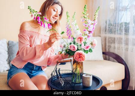Frau macht Strauß von Rosen und Füchshandschuhen Blumen in Korb in Vase zu Hause gepflückt. Einrichtung und Einrichtung im Sommer. Stockfoto