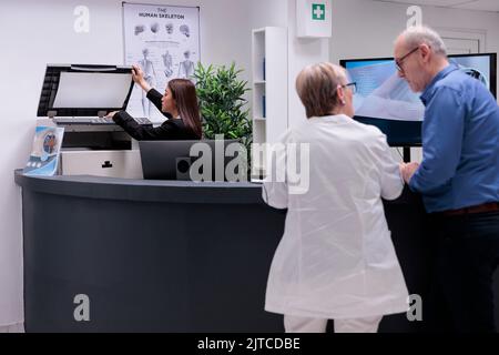 Mitarbeiter, der am Empfang einen Kopierdrucker verwendet, um vor der Untersuchung Berichte zu erstellen. Frau an der Rezeption, die mit Formularen am Schalter für die Registrierung an Gesundheitsterminen arbeitet. Stockfoto