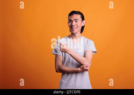 Fröhlich lächelnder asiatischer Mann, der zur Seite zeigt, mit Zeigefinger, Werbeprodukt. Teenager stehen mit positivem Gesichtsausdruck, zeigen Werbung, Studio Medium shot Stockfoto