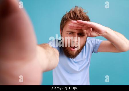 Porträt eines jungen erwachsenen, hübschen, neugierigen bärtigen Mannes, der mit aufmerksamer Sicht und Blick in die Ferne blickt und die Hand nahe an der Stirn hält. Innenaufnahme des Studios isoliert auf blauem Hintergrund. Stockfoto