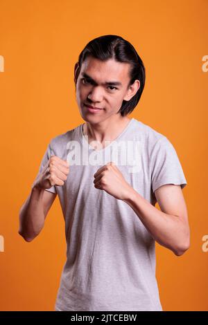 Wütender, starker junger asiatischer Mann, der in einer kämpfenden Pose mit ernstem Gesichtsausdruck steht. Wütender Teenager in Boxposition mit geballten Fäusten Porträt, aggressive Kämpfer Blick auf die Kamera Stockfoto