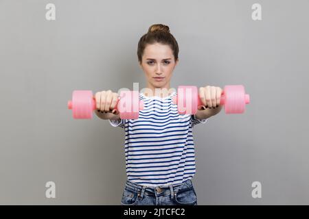 Porträt einer ernsthaft konzentrierten Frau, die ein gestreiftes T-Shirt trägt und rosa Hanteln an die Kamera hält und alleine arbeitet. Innenaufnahme des Studios isoliert auf grauem Hintergrund. Stockfoto