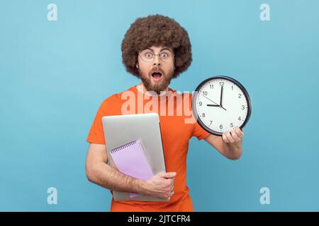 Porträt eines schockierten Mannes mit Afro-Frisur in orangefarbenem T-Shirt, der ein Notizbuch und einen Laptop in der Hand hält, mit großer Wanduhr und Deadline. Innenaufnahme des Studios isoliert auf blauem Hintergrund. Stockfoto