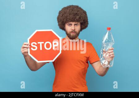 Porträt eines verantwortungsbewussten Mannes mit Afro-Frisur, das ein orangefarbenes T-Shirt mit rotem Stoppschild und einer Plastikflasche trägt und die Kamera anschaut. Innenaufnahme des Studios isoliert auf blauem Hintergrund. Stockfoto