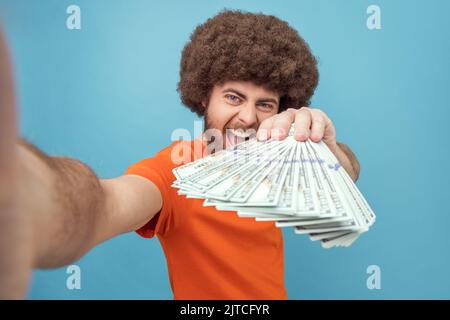 Porträt eines verrückten reichen Mannes mit Afro-Frisur in orangefarbenem T-Shirt, der den Abonnenten viele Dollar-Banknoten zeigt, mit aufgeregt Ausdruck, POV. Innenaufnahme des Studios isoliert auf blauem Hintergrund. Stockfoto