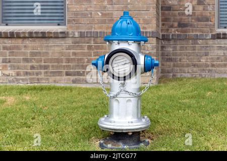 Grauer und blauer Hydrant auf dem Rasen auf dem Hof in einem Wohnkomplex mit Backsteinmauer des Gebäudes im Hintergrund Stockfoto