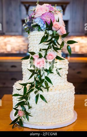 Weiße Hochzeitstorte mit kaskadierenden rosa und lila Blüten und Blättern auf einer Theke sitzen. Stockfoto