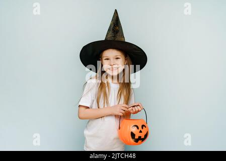 Ein kleines Mädchen in einem Hexenhut hält einen Süßigkeitenkorb auf blauem Hintergrund und lächelt. Trick or Treat. Stockfoto