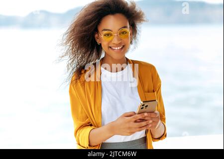 Das moderne, modische afroamerikanische Mädchen mit lockigem Haar, das ein orangefarbenes Hemd und eine orangefarbene Brille trägt, steht im Freien, nutzt ihr Smartphone, um Nachrichten mit Freunden auszutauschen, blickt auf die Kamera und lächelt Stockfoto