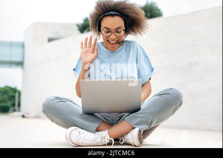 Clevere, glückliche, afroamerikanische junge Frau mit Headset, Angestellte des Unternehmens, führt Ferngespräche, verhandelt mit Kollegen, sitzt im Freien, mit Laptop, spricht über Videoanruf, lächelt Stockfoto