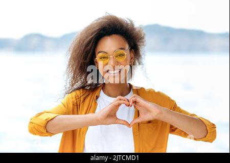 Foto von einem schönen positiv stilvolle lockige afroamerikanische Mädchen, mit oranger Brille, steht im Freien, zeigt eine Herzform mit ihren Händen, ein Zeichen der Liebe, Frieden, schaut auf die Kamera, lächelt Stockfoto