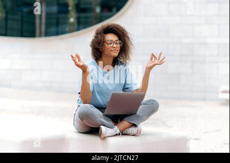 Ruhige, lockige, afroamerikanische junge Frau mit Brille, Büroangestellte, sitzt in einer Lotusposition im Freien in der Nähe des Geschäftszentrums, meditiert, entspannt, schließt ihre Augen. Pause während der Online-Arbeit Stockfoto