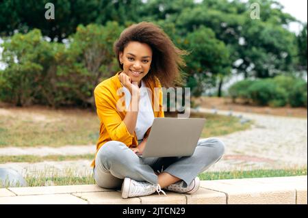 Lovely positive clever afroamerikanischen lockigen tausendjährigen Mädchen, Studentin, Freiberufler, in stilvollen Freizeitkleidung, sitzen mit einem Laptop im Freien, Blick auf die Kamera, lächelnd glücklich Stockfoto