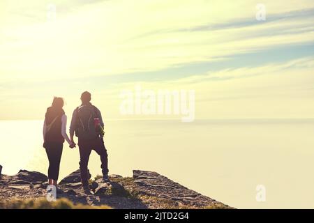 Nehmen Sie sich nur Erinnerungen, hinterlassen Sie nur Fußabdrücke. Rückansicht eines jungen Paares, das die Aussicht von der Spitze eines Berges bewundert. Stockfoto