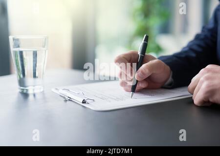 Ein Geschäftsmann, der Papierkram auf einem Klemmbrett ausfüllt, während er an einem Tisch in einem Büro sitzt. Stockfoto