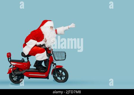 In voller Länge ein Seitenansicht-Foto eines Santaklaus, der mit einem roten Elektromoped fährt, dessen Arm nach vorne gestreckt ist, in einer Superman-Pose auf einem blau isolierten Hintergro Stockfoto