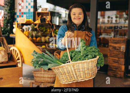 Supermarktmitarbeiter mit Down-Syndrom hält einen Korb mit frischem Bio-Gemüse in einem Lebensmittelgeschäft. Glückliche Frau mit einer intellektuellen Behinderung w Stockfoto