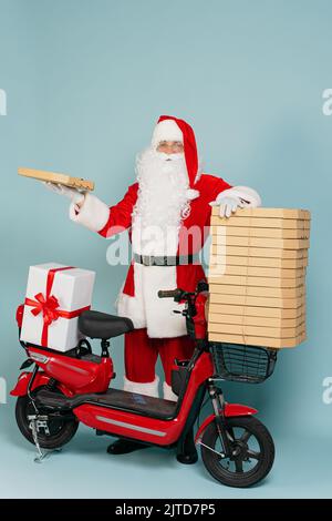 Der weihnachtsmann steht in vollem Wachstum auf einem riesigen Stapel von Pizzabällen und hält eine Schachtel in der Hand vor dem Hintergrund eines roten Elektromopeds auf einem Stockfoto