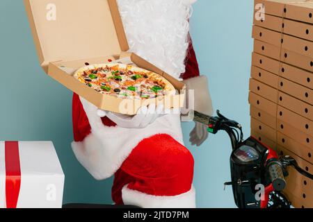 Nahaufnahme einer offenen Schachtel Pizza in den Händen eines Lieferers in einem weihnachtsmann-Kostüm. Er hält sich in den Händen vor dem Hintergrund eines roten Mopeds mit einem Stockfoto