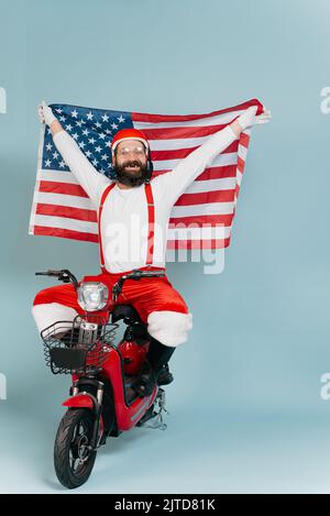 Ein Hipster im santa-Kostüm hält die usa-Flagge in der Hand über seinen Kopf, während er auf einem roten Elektroroller auf einem blau isolierten Hintergrund mit c sitzt Stockfoto