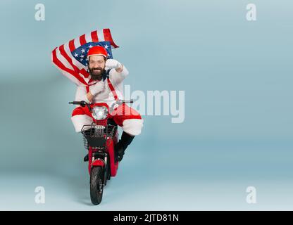 Santa in einer Superman-Pose mit einer flatternden usa-Flagge in Form eines Umhangs und einem Helm fährt ein roter Elektroroller auf einem blau isolierten Hintergrund mit Stockfoto