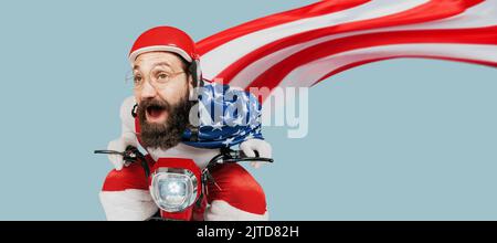 Der Weihnachtsmann mit einer großen us-Flagge in Form eines Regenmantels fährt mit hoher Geschwindigkeit auf einem Elektroroller in Richtung Kamera. Junge Erwachsene Hipster in einem roten Helm Stockfoto