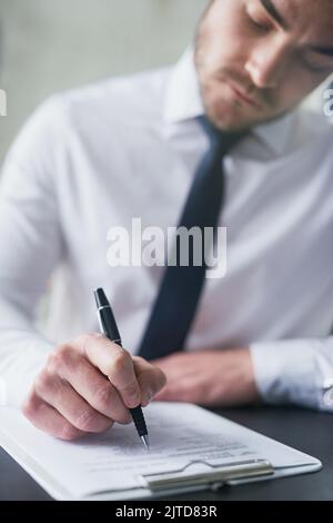 HES gut vorbereitet für diesen Fragebogen. Ein junger Geschäftsmann, der Papierkram auf einem Klemmbrett ausfüllt, während er an einem Tisch in einem Büro sitzt. Stockfoto