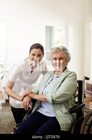 Sie bringt ihren Patienten immer ein Lächeln. Porträt einer lächelnden Betreuerin und einer älteren Frau im Rollstuhl zu Hause. Stockfoto