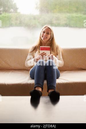 Es gibt nichts ganz wie ein gutes Buch. Porträt einer attraktiven jungen Frau, die ein Buch in der Hand hält, während sie zu Hause auf dem Sofa sitzt. Stockfoto