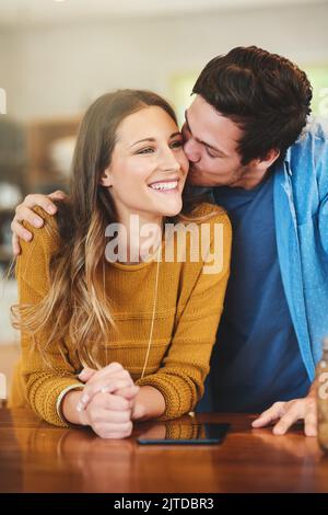 Verbreiten Sie die Liebe. Ein liebevoller junger Mann küsst seine Freundin auf die Wange in ihrer Küche. Stockfoto