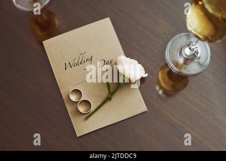 Draufsicht der Hochzeitskomposition auf dem Tisch, bestehend aus Einladung, frischer weißer Rose und zwei Ringen, die von Sektflöten umgeben sind Stockfoto