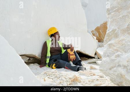Junge, erholsame Arbeiterin in Uniform und Schutzhelm, die nach der Arbeit Pause gemacht hat, während sie am Steinbruch gegen riesigen Marmorfelsen sitzt Stockfoto