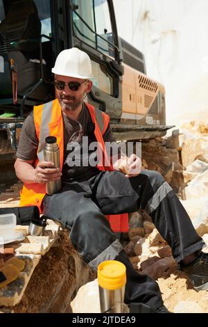 Männlicher Ingenieur mittleren Alters in Arbeitskleidung und Harthut mit Sandwich und heißem Tee zum Mittagessen, während er auf der Raupe des Baggers sitzt Stockfoto