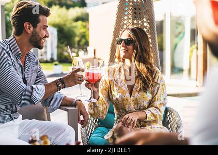 Ein junges Paar in der Generation der Millennials feiert einen Toast, der im Garten des Resorts mit klinkenden Gläsern bei fantasievollen Cocktails sitzt - Erholung im Hinterhof Stockfoto