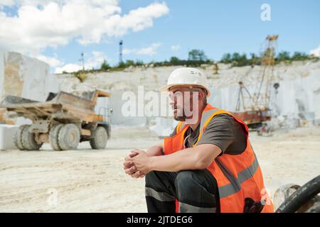 Müder Arbeiter des Marmorsteinbruchs, der am Arbeitsplatz vor der Kamera sitzt, gegen einen Muldenkipper und einen funktionierenden Bulldozer mit einem Kran Stockfoto