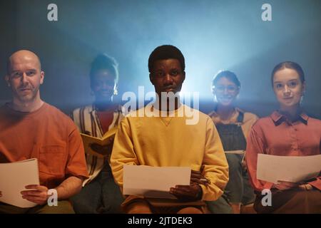 Die Moody-Aufnahme eines schwarzen jungen Mannes, der von Projektorstrahlen beleuchtet wird, sitzt auf einem Stuhl und wird bei einem Geschäftsseminar von den Zuschauern aufgenommen Stockfoto