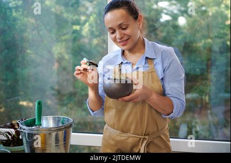 Charmante Gärtnerin trägt eine beige Schürze auf der Veranda zu Hause, Pflanzen in neue Keramik-Töpfe zu verpflanzen. Gartenarbeit. Stockfoto