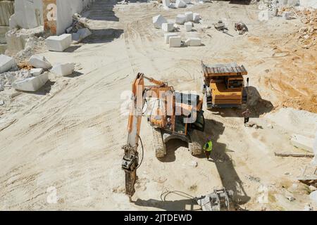 Über dem Winkel des Muldenkipper und der hydraulischen Raupenbaumaschine mit Grabber, der auf dem Gebiet des riesigen Marmorbruchs arbeitet Stockfoto