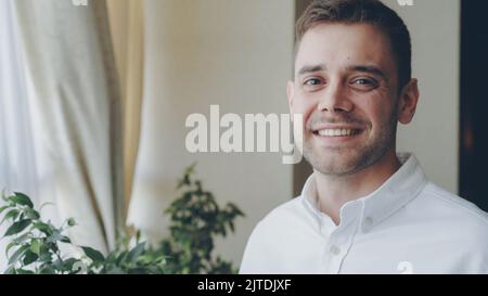 Nahaufnahme eines dunkelhaarigen, hübschen jungen Mannes in weißem Hemd, der fröhlich lächelt und die Kamera anschaut. Junge Menschen, schöne Inneneinrichtung und Glückskonzept. Stockfoto