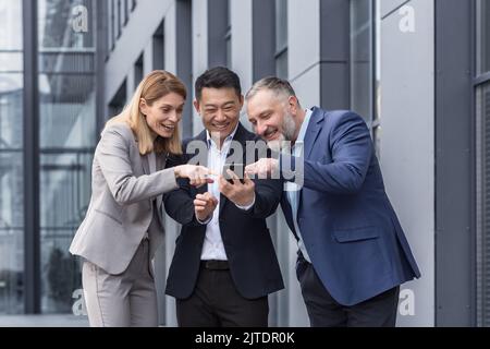 Vielfältige Geschäftsgruppe, drei männliche und weibliche Mitarbeiter vor dem Bürogebäude, fröhlich lächelnd und glücklich beim Ansehen von Videos am Telefon Stockfoto