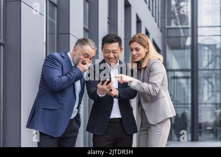 Vielfältige Geschäftsgruppe, drei männliche und weibliche Mitarbeiter vor dem Bürogebäude, fröhlich lächelnd und glücklich beim Ansehen von Videos am Telefon Stockfoto