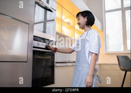 Junge Hausfrau, die etwas im Ofen backt und beschäftigt aussieht Stockfoto