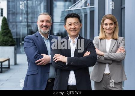 Erfolgreiches, vielfältiges Geschäftsteam, drei lächelnde und vor die Kamera schauende Mitarbeiter, Traumteam mit asiatischem Chef vor dem Bürogebäude, Kollegen in Geschäftsanzügen Stockfoto